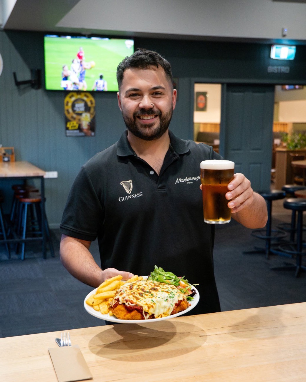 A member of the Newborough Hotel team holding a chicken parma and a cold beer in the bistro